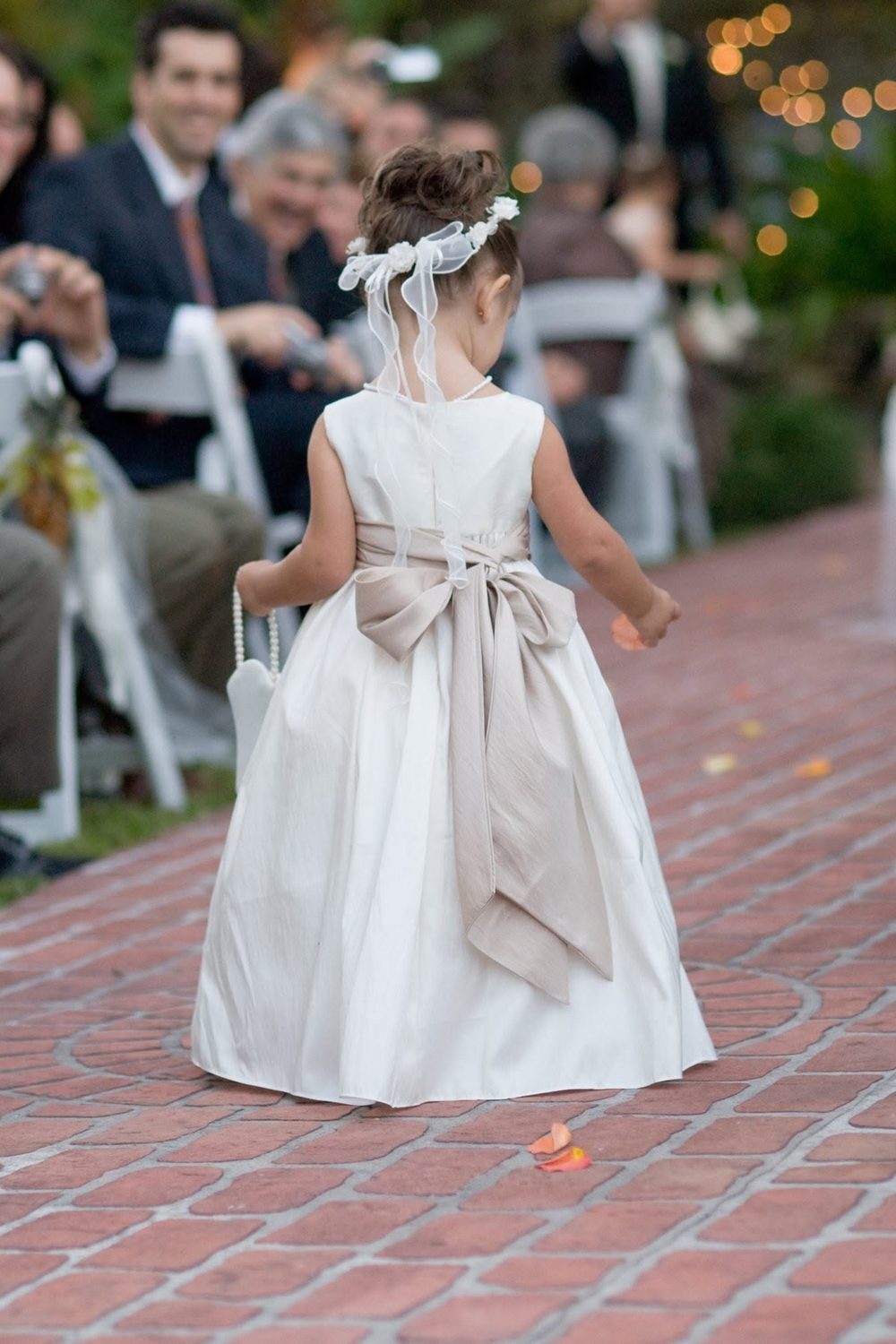 A flower girl in an ivory dress walking a brick aisle past seated guests at an outdoor ceremony