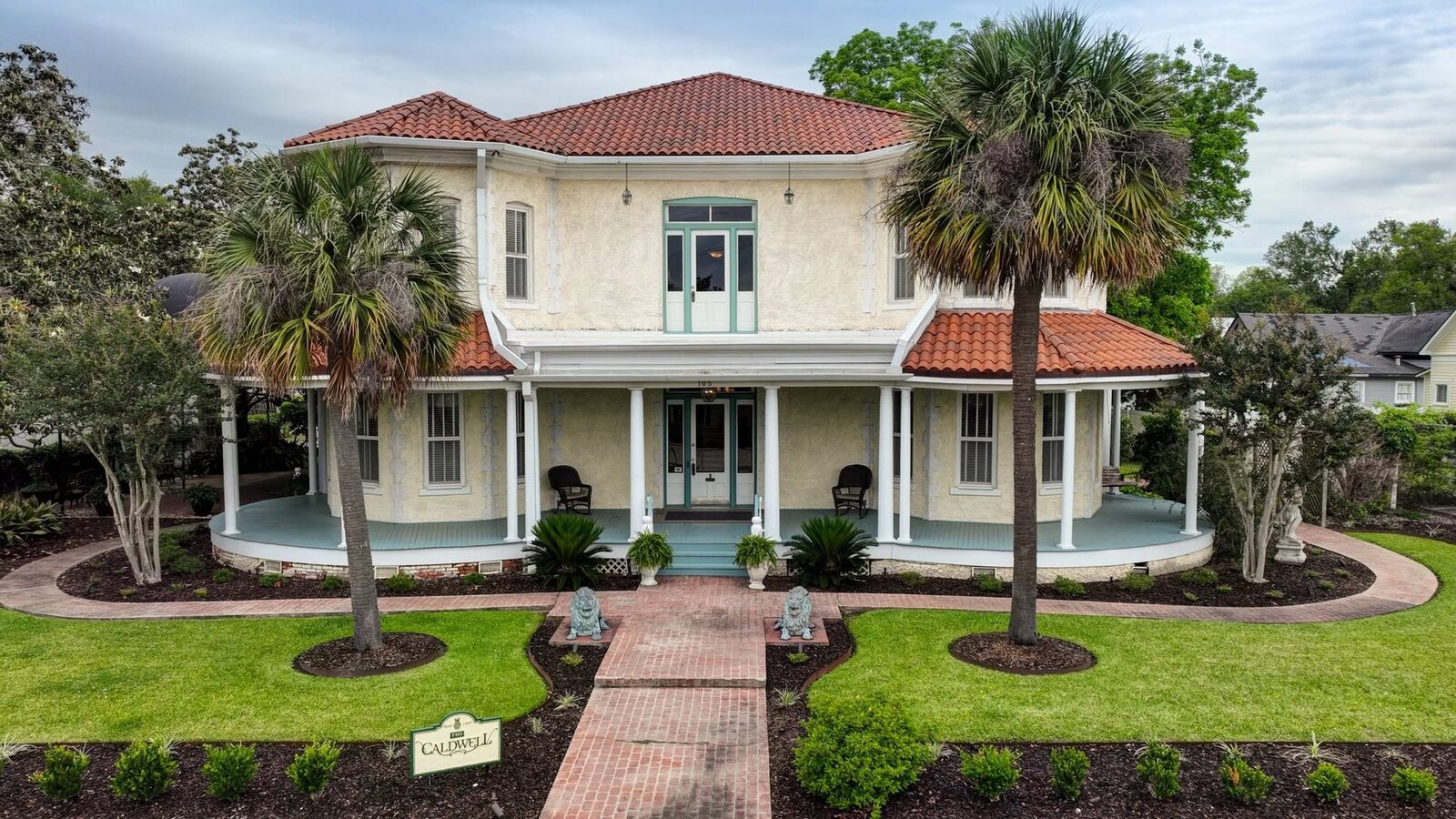 The Caldwell House, a historic home with a curved wraparound porch, terra-cotta roof, and palm-lined brick walk