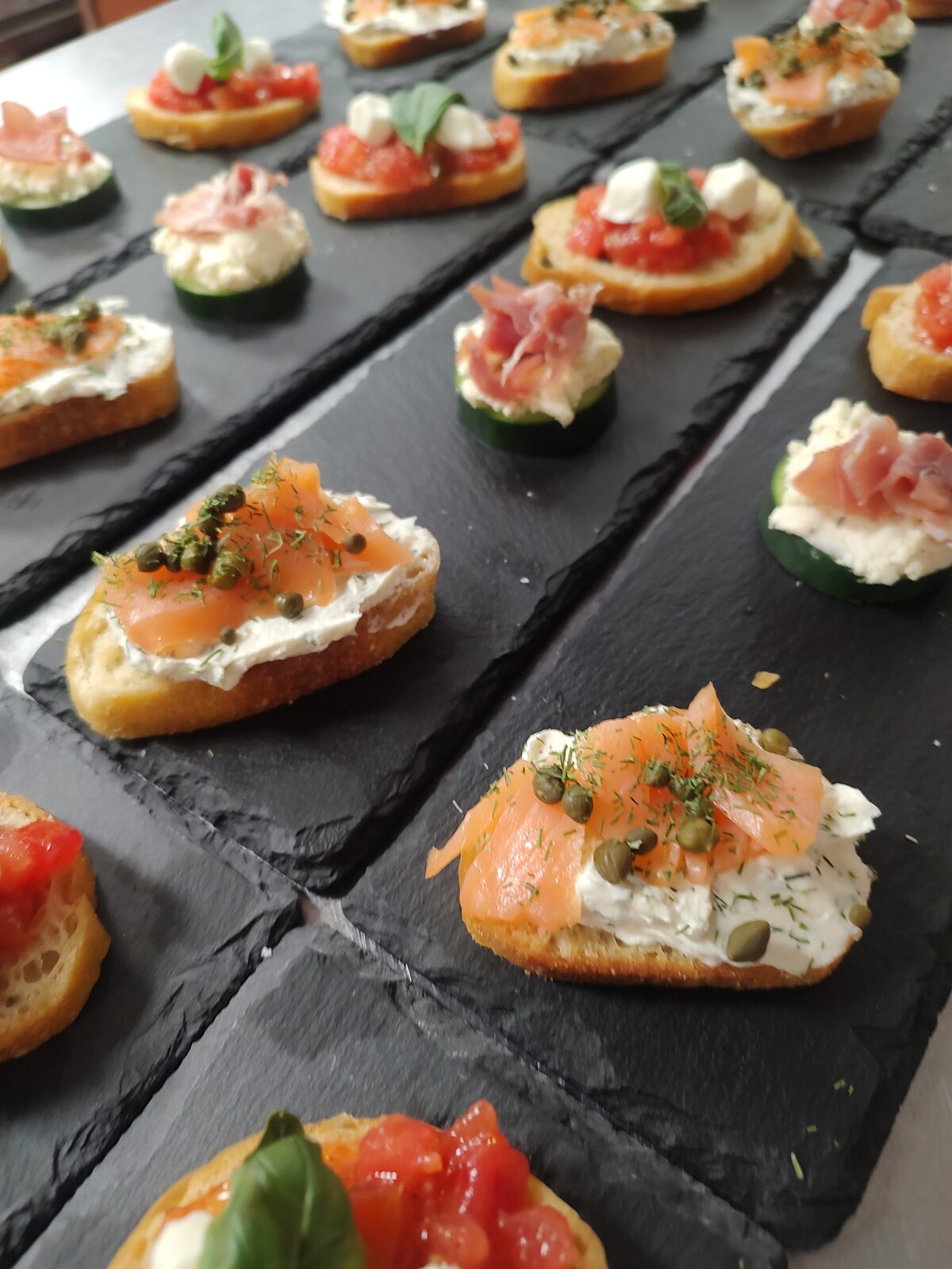 Rows of canapés on slate — smoked salmon with dill and capers, bruschetta with basil, and prosciutto on cucumber