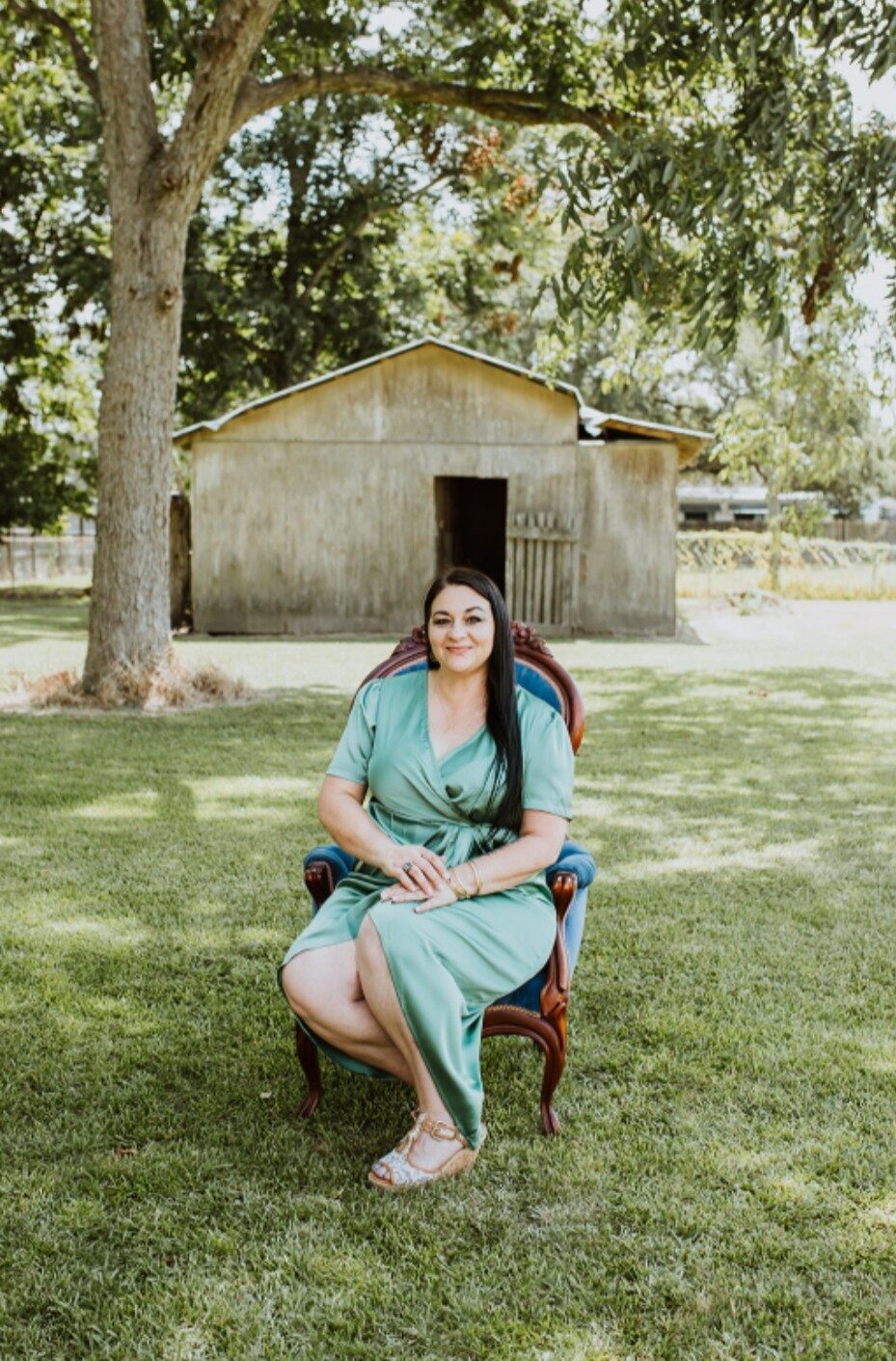 Jill Bernard, founder of The Southern Spread, seated in a vintage blue velvet chair beneath a pecan tree in a sage green dress