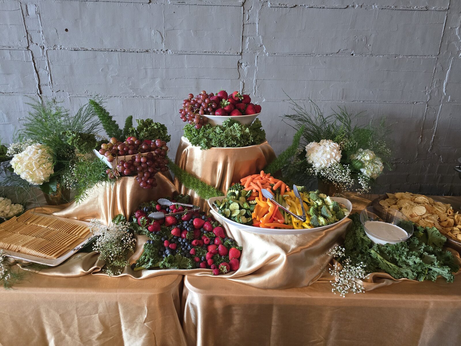 A grazing table draped in gold satin — tiered bowls of strawberries and grapes, fresh berries, garden vegetables, and white hydrangeas