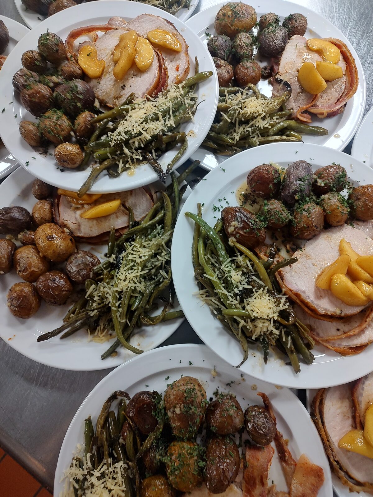 Plated suppers ready for service — bacon-wrapped roasted pork loin with spiced apples, rosemary roasted potatoes, and parmesan green beans