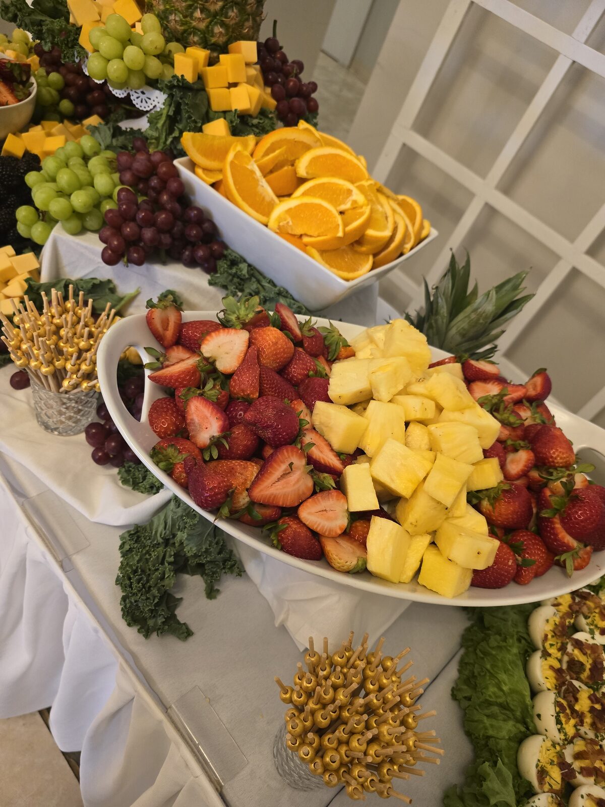 A fruit display of strawberries, pineapple, and orange slices nestled in kale, with cheese, gold picks, and trays of deviled eggs beyond