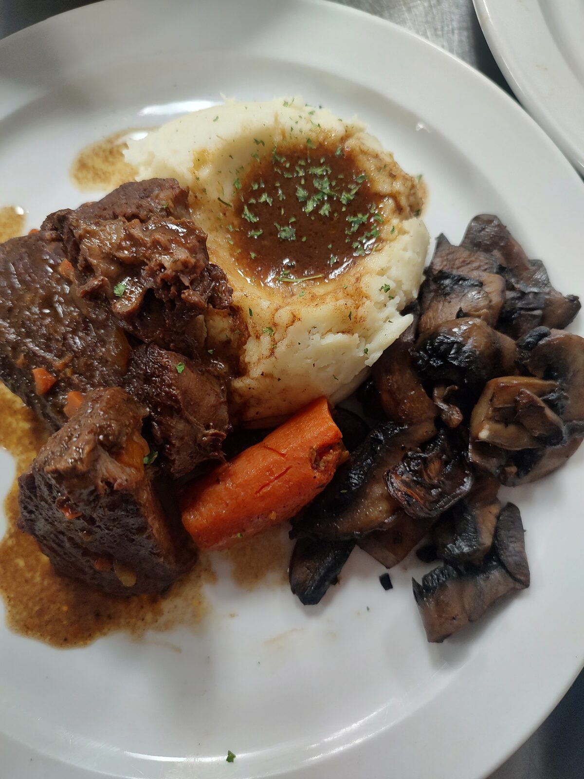 Braised beef short ribs plated with mashed potatoes and gravy, sautéed mushrooms, and a glazed carrot